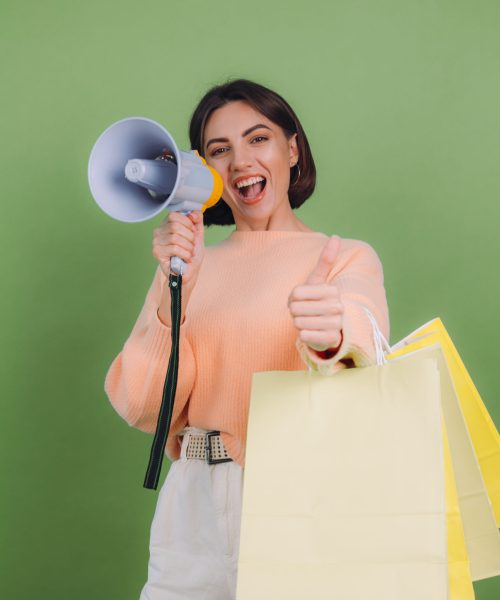 Young woman in casual peach sweater isolated on green olive color background shout in megaphone holding shopping bags, announces discounts sale promotion
